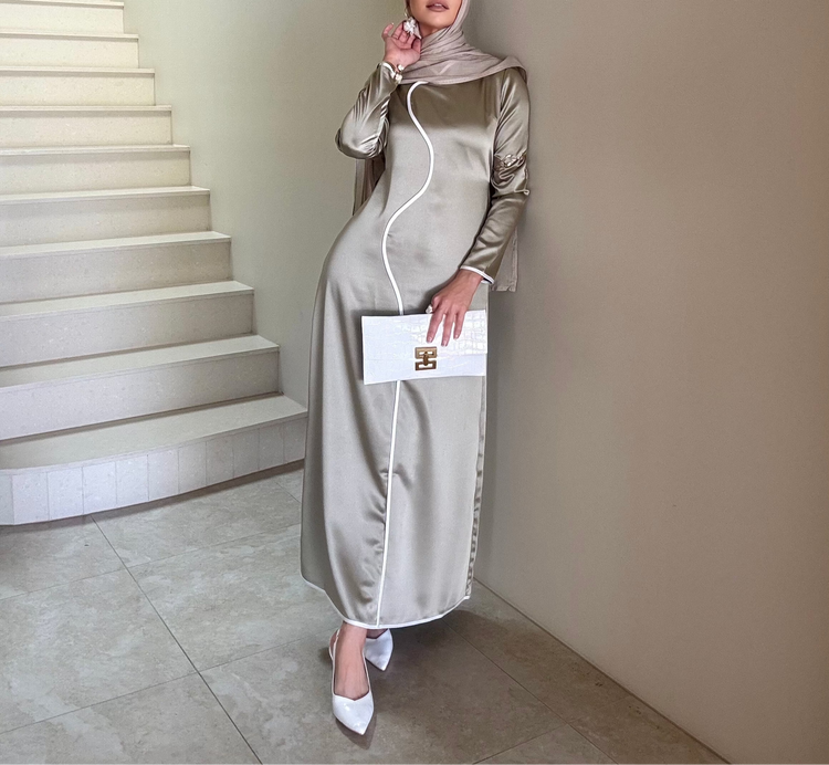 Woman in an olive satin dress with white trim leaning against a wall by a staircase