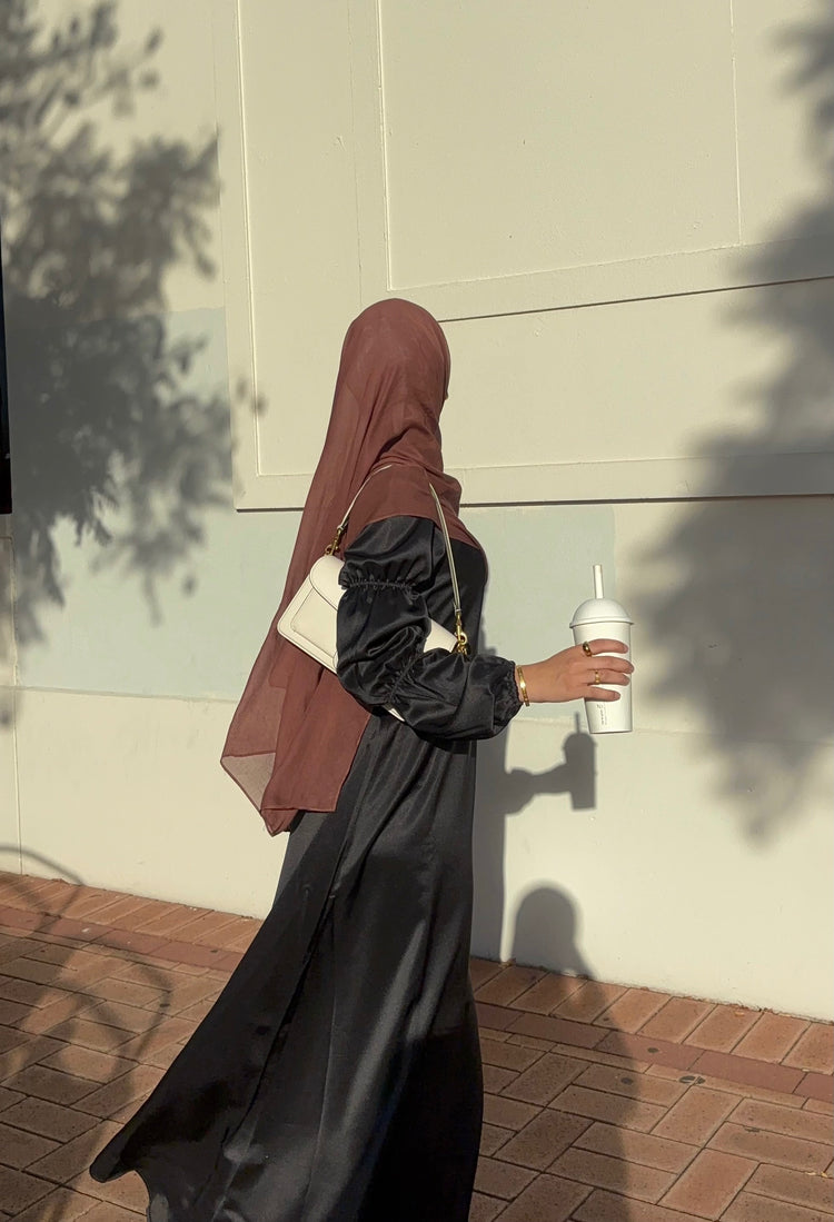 Woman in a black satin dress with puff sleeves walking past a sunlit wall